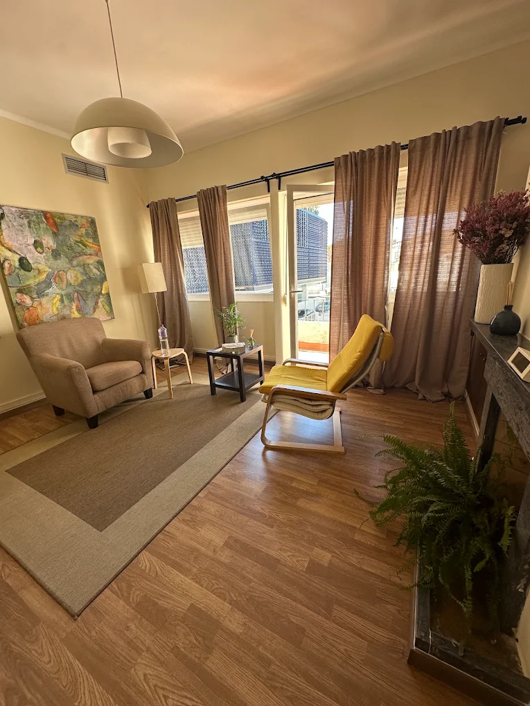 Sala de consulta com poltrona bege, cadeira amarela, cortinas de linho, plantas e um quadro abstrato